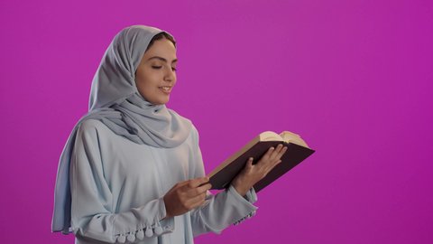 إيماءات التركيز والاندماج ، ثقافة التعلم الذاتي، بورتريه لامرأة عربية خليجية سعودية ترتدي العباءة الرمادية والحجاب تحمل بيدها كتاب تثقيفي, الاستمتاع بقراءة الروايات والقصص, خلفية بنفسجية