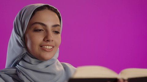 ثقافة التعلم الذاتي، الاستمتاع بقراءة الروايات والقصص , إيماءات التركيز والاندماج ، بورتريه لامرأة عربية خليجية سعودية ترتدي العباءة الرمادية والحجاب تحمل بيدها كتاب تثقيفي, خلفية بنفسجية