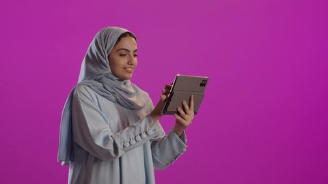 Using modern and advanced technology, a portrait of a veiled Saudi Arabian woman wearing a gray abaya, using a tablet and browsing social media, against a purple background.