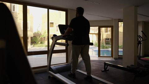 Using sports equipment, practicing warm-up exercises, maintaining health and physical fitness, a young Arab Gulf Saudi man wearing sports clothes walking on the treadmill, sports club, engaging in enjoyable recreational activities.