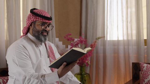 ثقافة التعلم الذاتي، تنمية مهارات القراءة,  قضاء أوقات الفراغ بالتثقيف والتعلم, الاهتمام بتصفح الكتب المفيدة، رجل عربي خليجي سعودي يرتدي الثوب التقليدي والشماغ يجلس على آريكة في المنزل ويحمل بيده كتاب، منزلتراثي بتصميم عصري، القراءة والمطالعة عادة يومية