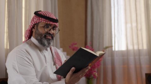 القراءة والمطالعة عادة يومية، ثقافة التعلم الذاتي، تنمية مهارات القراءة,  قضاء أوقات الفراغ بالتثقيف والتعلم, الاهتمام بتصفح الكتب المفيدة، رجل عربي خليجي سعودي يرتدي الثوب التقليدي والشماغ يجلس على آريكة في المنزل ويحمل بيده كتاب، منزلتراثي بتصميم عصري