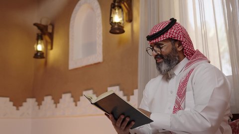 التصفح وتقليب الصفحات، القراءة والمطالعة عادة يومية، تنمية مهارات القراءة,  قضاء أوقات الفراغ بالتثقيف والتعلم, الاهتمام بتصفح الكتب المفيدة، رجل عربي خليجي سعودي يرتدي الثوب التقليدي والشماغ يجلس على آريكة في المنزل ويحمل بيده كتاب، منزلتراثي بتصميم عصري