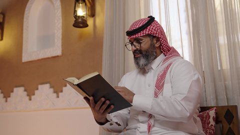 استخدام النظارة الطبية اثناء القراءة، المطالعة عادة يومية، تنمية مهارات القراءة,  قضاء أوقات الفراغ بالتثقيف والتعلم, الاهتمام بتصفح الكتب المفيدة، رجل عربي خليجي سعودي يرتدي الثوب التقليدي والشماغ يجلس على آريكة في المنزل ويحمل بيده كتاب، منزلتراثي بتصميم عصري