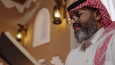 تعابير الاندماج والتركيز مع الكتاب، المطالعة عادة يومية، تنمية مهارات القراءة,  قضاء أوقات الفراغ بالتثقيف والتعلم, الاهتمام بتصفح الكتب المفيدة، رجل عربي خليجي سعودي يرتدي الثوب التقليدي والشماغ يجلس على آريكة في المنزل ويحمل بيده كتاب، منزلتراثي بتصميم عصري