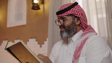 تنمية مهارات القراءة,  قضاء أوقات الفراغ بالتثقيف والتعلم, الاهتمام بتصفح الكتب المفيدة، رجل عربي خليجي سعودي يرتدي الثوب التقليدي والشماغ يجلس على آريكة في المنزل ويحمل بيده كتاب، منزلتراثي بتصميم عصري، ثقافة التعلم الذاتي، القراءة والمطالعة عادة يومية