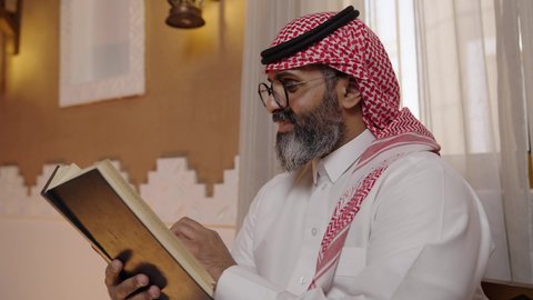 قضاء أوقات الفراغ بالتثقيف والتعلم, تنمية مهارات القراءة, الاهتمام بتصفح الكتب المفيدة، رجل عربي خليجي سعودي يرتدي الثوب التقليدي والشماغ يجلس على آريكة في المنزل ويحمل بيده كتاب، منزلتراثي بتصميم عصري، ثقافة التعلم الذاتي، القراءة والمطالعة عادة يومية