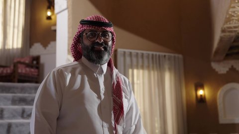 Gestures of joy and happiness, attention to appearance, looking at the camera with gestures of pride and dignity, the concept of masculinity and elegance, a man inside a modern home, a photo of a Saudi Arabian Gulf gentleman wearing a white thobe and a ghutrah walking between houses, wearing glasses on his eyes.