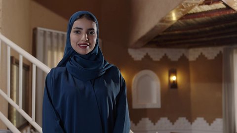 الاهتمام بالمظهر الخارجي، مفهوم الانوثة والنعومة، إيماءات السرور والسعادة، سيدة عربية خليجية سعودية محجبة مبتسمة ترتدي عباءة والحجاب تقف داخل منزل تراثي عصري، توثيق اللحظات السعيدة، استخدام السلالم داخل المنزل للتنقل بسهولة