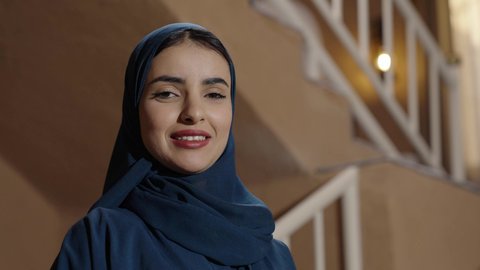 مفهوم الانوثة والنعومة، الاهتمام بالمظهر الخارجي، إيماءات السرور والسعادة، سيدة عربية خليجية سعودية محجبة مبتسمة ترتدي عباءة والحجاب تقف داخل منزل تراثي عصري، توثيق اللحظات السعيدة، استخدام السلالم داخل المنزل للتنقل بسهولة