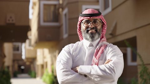 الوقوف بايدي متقاطعة، النظر إلى الكاميرا بايماءات بفخر واعتزاز ، مفهوم الرجولة والأناقة، رجل عند مدخل منزل عصري ، تصوير من الخلف سيد عربي خليجي سعودي يرتدي الثوب الابيض والشماغ يسير بين المنازل، نظارة طبية على عينيه، الاهتمام بالمظهر الخارجي