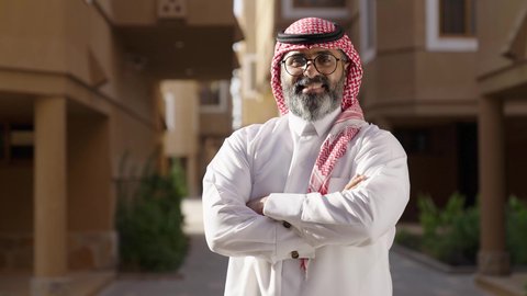 Attention to appearance, standing with crossed arms, looking at the camera with gestures of pride and dignity, the concept of masculinity and elegance, a man at the entrance of a modern house, photographed from behind, an Arab Gulf Saudi man wearing a white thobe and a ghutrah walking between houses, wearing glasses on his eyes.