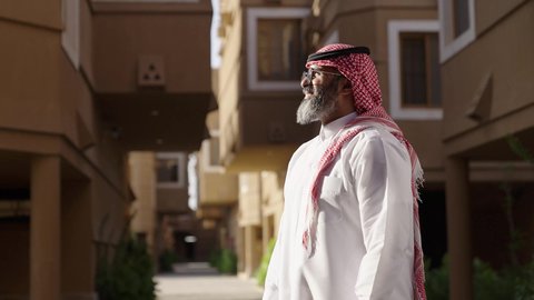 النظر إلى الكاميرا بايماءات بفخر واعتزاز ، مفهوم الرجولة والأناقة، رجل عند مدخل منزل عصري ، التأمل والتعمق في تفاصيل المبنى الخارجي للمنزل، تصوير لسيد عربي خليجي سعودي يرتدي الثوب الابيض والشماغ يسير بين المنازل، نظارة طبية على عينيه، الاهتمام بالمظهر الخارجي