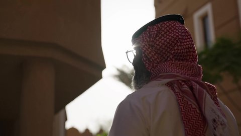 The concept of masculinity and elegance, a man at the entrance of a modern house, contemplating and delving into the details of the house's exterior, a shot from behind of an Arab Gulf Saudi man wearing a white thobe and a ghutrah walking between the houses, wearing glasses on his eyes, paying attention to his appearance, standing with pride and dignity.