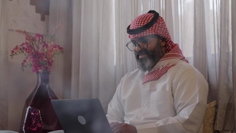 استخدام التقنية الحديثه المتطورة في الحياة العملية، العمل عن بعد، مهنة ووظيفة مكتبية، ايماءات الاندماج والتركيز، رجل عربي خليجي سعودي يرتدي الثوب التقليديوالشماغ ويضع النظارة الطبية يجلس على الكرسي ويعمل على الحاسب المحمول في صالة المنزل، النظر الى شي ما