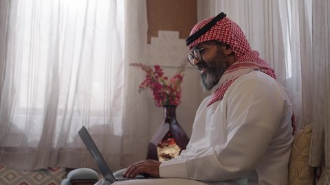 إنجاز المهام في المنزل، استخدام التقنية الحديثه المتطورة في الحياة العملية، مهنة ووظيفة مكتبية، ايماءات الاندماج والتركيز، رجل عربي خليجي سعودي يرتدي الثوب التقليديوالشماغ ويضع النظارة الطبية يجلس على الكرسي ويعمل على الحاسب المحمول في صالة المنزل، النظر الى شي ما