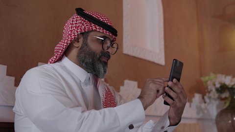 استخدام التكنولوجيا والأجهزة التقنية، التسوق الالكتروني وتصفح برامج وتطبيقات الجوال، استخدام الهاتف المحمول، تصوير لرجل عربي خليجي سعودي يرتدي الثوب التقليدي والشماغ  والنظارات الطبية يجلس في صالة المنزل التراثي بتصميم عصري يحمل بيده الهاتف المحمول