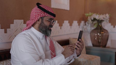 انهاء المهام عن بعد، استخدام التكنولوجيا والأجهزة التقنية، التسوق الالكتروني وتصفح برامج وتطبيقات الجوال، تصوير لرجل عربي خليجي سعودي يرتدي الثوب التقليدي والشماغ  والنظارات الطبية يجلس في صالة المنزل التراثي بتصميم عصري يحمل بيده الهاتف المحمول