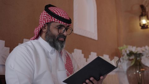 القراءة والمطالعة عادة يومية، قضاء أوقات الفراغ بالتثقيف والتعلم, تنمية مهارات القراءة, الاهتمام بتصفح الكتب المفيدة، رجل عربي خليجي سعودي يرتدي الثوب التقليدي والشماغ يجلس على آريكة في المنزل ويحمل بيده كتاب، منزلتراثي بتصميم عصري، ثقافة التعلم الذاتي