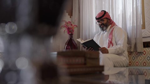 مفهوم الثقافة والتعلم، الاستمتاع في أوقات الفراغ بالتثقيف والتعلم, القراءة والمطالعة عادة يومية، الاهتمام بتصفح الكتب المفيدة، رجل عربي خليجي سعودي يرتدي الثوب التقليدي والشماغ يجلس على آريكة في المنزل ويحمل بيده كتاب، منزلتراثي بتصميم عصري، ثقافة التعلم الذاتي