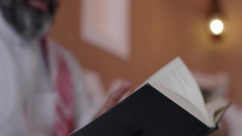 الاستمتاع في أوقات الفراغ بالتثقيف والتعلم, القراءة والمطالعة عادة يومية، تنمية مهارات القراءة, الاهتمام بتصفح الكتب المفيدة، رجل عربي خليجي سعودي يرتدي الثوب التقليدي والشماغ يجلس على آريكة في المنزل ويحمل بيده كتاب، منزلتراثي بتصميم عصري، ثقافة التعلم الذاتي