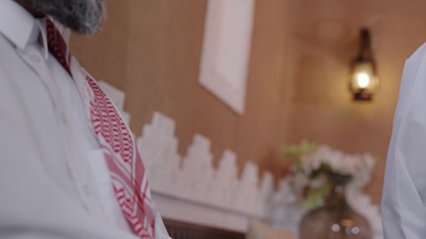 التسامر وتبادل الاحاديث، تعابير المودة والمحبة، دور الأب في حياه الأبناء،  الاستمتاع بقضاء وقت مع الابناء، رجل كبير بالسن عربي خليجي سعودي يرتدي الثوب التقليدي والشماغ يجلس على الاريكة بجانب ابنه  في غرفة الجلوس، جلسة عائلية سعيدة