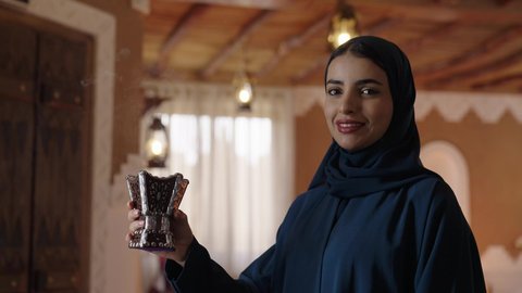 The beauty of Saudi women, the concept of femininity, elegance, chic, and softness; attention to outward appearance; modern and contemporary hijab fashion; a close-up photograph of a veiled Saudi Arabian woman wearing a hijab and abaya, perfuming herself using an incense burner; distinctive Gulf scents and perfumes.