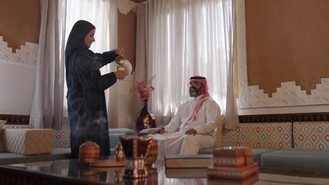 تقديم المشروبات الساخنة العربية ،كرم الضيافة وحسن الاستقبال في السعودية, أصالة عادات وتقاليد المجتمع السعودي في المناسبات الاسلامية,  إمرأة عربية خليجية سعودية مبتسمة ترتدي العباءة والحجاب تقوم بصب القهوة الخليجية وتقديمها للضيوف في صالة المنزل