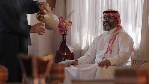 صلة الرحم والترابط الاسري، كرم الضيافة وحسن الاستقبال في السعودية, أصالة عادات وتقاليد المجتمع السعودي في المناسبات الاسلامية, إمرأة عربية خليجية سعودية مبتسمة ترتدي العباءة والحجاب تقوم بصب القهوة الخليجية وتقديمها للضيوف في صالة المنزل