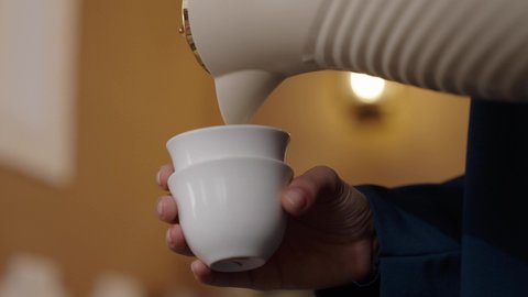 Pouring coffee from a copper pot: a symbol of Saudi hospitality and warm welcome, reflecting the authentic customs and traditions of Saudi society during Islamic occasions. A close-up photograph shows the hands of a smiling Saudi Arabian woman, wearing an abaya and hijab, pouring and serving traditional Gulf coffee to guests in the living room.
