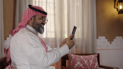 التسوق الالكتروني وتصفح برامج وتطبيقات الجوال، تطور التكنولوجيا والأجهزة التقنية،  استخدام الهاتف المحمول، تصوير لرجل عربي خليجي سعودي يرتدي الثوب التقليدي والشماغ  والنظارات الطبية يجلس في صالة المنزل التراثي بتصميم عصري يحمل بيده الهاتف المحمول
