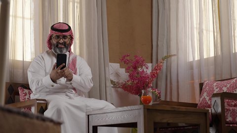 تطور التكنولوجيا والأجهزة التقنية،  استخدام الهاتف المحمول،  التسوق الالكتروني وتصفح برامج وتطبيقات الجوال، تصوير لرجل عربي خليجي سعودي يرتدي الثوب التقليدي والشماغ  والنظارات الطبية يجلس في صالة المنزل التراثي بتصميم عصري يحمل بيده الهاتف المحمول