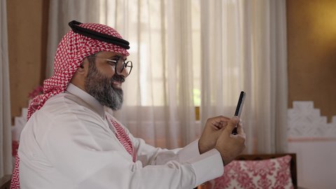 Mobile phone use, technological advancements and devices, e-commerce and browsing mobile applications: A photograph of a Saudi Arabian Gulf Arab man wearing a traditional thobe, shemagh, and eyeglasses, sitting in a modernly designed heritage home, holding a mobile phone.