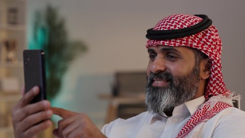 تصوير مقرب لرجل عربي خليجي سعودي يرتدى الثوب والشماغ يمسك بيده الهاتف المحمول,الدردشة مع الأهل والأصدقاء, إستخدام أجهزة تقنية حديثة, ، دمج التقنية والتكنولوجيا بالحياة اليومي