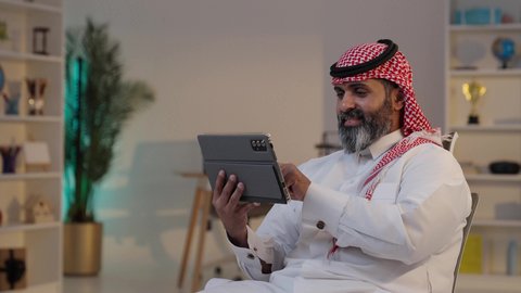 ايماءات وتعابير تدل على السعادة, رجل عربي خليجي سعودي يرتدى الثوب والشماغ يجلس على الكرسى يستخدم التابلت ويقوم بتصفح وسائل التواصل الإجتماعى, استخدام التكنولوجيا الحديثة