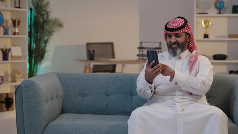 Enjoying quality time on his mobile phone with friends; a Saudi Arabian man wearing a thobe and shemagh uses his mobile phone, online shopping, and browsing social media.