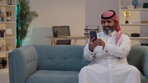انجاز المهام عن بعد, تصفح تطبيقات الهاتف المحمول, استخدام جهاز تقني حديث، دمج التكنولوجيا في الحياة اليومية, رجل عربي خليجي سعودي يرتدى الثوب والشماغ يستخددم الجوال