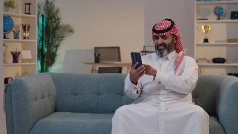 Browse mobile apps, use modern technology, accomplish tasks remotely, integrate technology into daily life: a Saudi Arabian Gulf Arab man wearing a thobe and shemagh using a mobile phone.