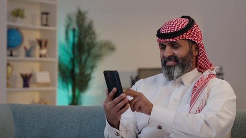 ايماءات وتعابير تدل على السعادة, استخدام التكنولوجيا الحديثة,  تصوير لرجل عربي خليجي سعودي يرتدى الثوب والشماغ يمسك بيده الجوال ,الجلوس على الاريكة, تصفح تطبيقات الهاتف المحمول