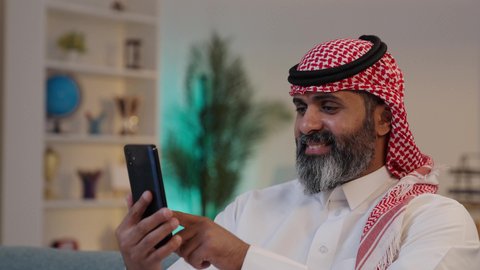 إستخدام الهاتف المحمول, ايماءات وتعابير تدل على السعادة, استخدام التكنولوجيا الحديثة,  تصوير من الجانب لرجل عربي خليجي سعودي يرتدى الثوب والشماغ يمسك بيده الجوال ,الجلوس على الاريكة, تصفح تطبيقات الهاتف المحمول