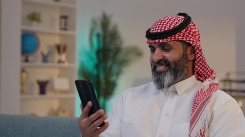 النظر إلى الجوال ومتابعة آخر الاخبار بإيماءات السعادة,  تصويرلرجل عربي خليجي سعودي يرتدى الثوب والشماغ يمسك بيده الجوال ,الجلوس على الاريكة, تصفح تطبيقات الهاتف المحمول, استخدام جهاز تقني حديث، دمج التكنولوجيا في الحياة اليومية