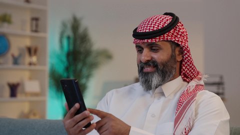 دمج التقنية والتكنولوجيا بالحياة اليومية,  تصوير لرجل عربي خليجي سعودي يرتدى الثوب والشماغ يمسك بيده الجوال ,الجلوس على الاريكة, تصفح تطبيقات الهاتف المحمول, استخدام جهاز تقني حديث