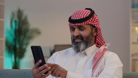 تصفح مواقع التواصل الاجتماعي,  تصويرلرجل عربي خليجي سعودي يرتدى الثوب والشماغ يمسك بيده الجوال ,الجلوس على الاريكة, تصفح تطبيقات الهاتف المحمول, استخدام جهاز تقني حديث، دمج التكنولوجيا في الحياة اليومية