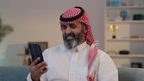 التكنولوجيا والتقنية الحديثة,  تصوير لرجل عربي خليجي سعودي يرتدى الثوب والشماغ يمسك بيده الجوال ,الجلوس على الاريكة, تصفح تطبيقات الهاتف المحمول, استخدام جهاز تقني حديث