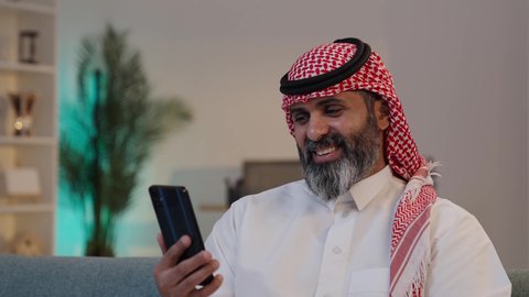 التواصل والاطمئنان عن الأهل والأصدقاء, تصويرلرجل عربي خليجي سعودي يرتدى الثوب والشماغ يمسك بيده الجوال ,الجلوس على الاريكة, تصفح تطبيقات الهاتف المحمول, استخدام جهاز تقني حديث، دمج التكنولوجيا في الحياة اليومية
