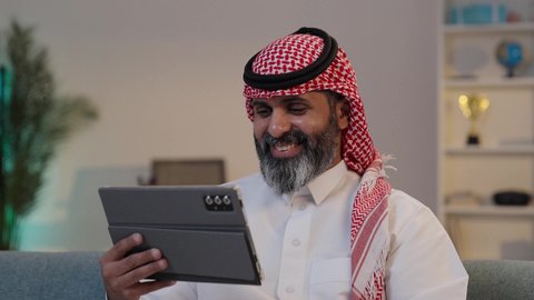 التواصل و الدردشة مع الاصدقاء بإيماءات السعادة، تصوير مقرب لرجل عربي خليجي سعودي يرتدى الثوب والشماغ يقوم بالنقرعلى جهاز التابلت, التسوق الإلكتروني وتصفح برامج وتطبيقات التواصل الاجتماع