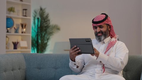 Online shopping and browsing social media apps; technological advancements and devices; a close-up photograph of a Saudi Arabian man wearing a thobe and shemagh, tapping on a tablet; communicating and chatting with family and friends.