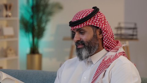 الشغف في ممارسة هواية المطالعة, استغلال فترات الراحة في التعلم وتنمية الفكر, الاهتمام بتصفح الكتب المفيدة, تصوير مقرب لرجل عربي خليجي سعودي يرتدى الثوب والشماغ يجلس على الاريكة يمسك بيده الكتاب
