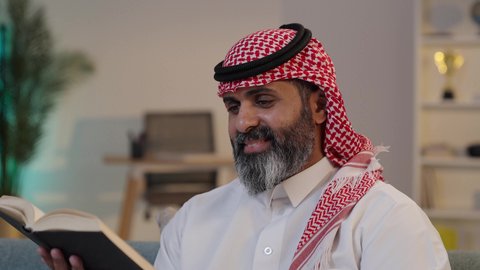The concept of contemplation and reflection, spending leisure time educating and learning, showing interest in browsing useful books, a close-up photograph of a Saudi Arabian Gulf man wearing a thobe and shemagh, sitting on a sofa holding a book, a love of reading.