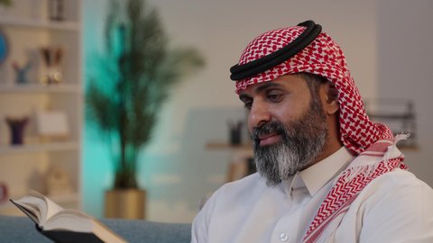 Utilizing breaks for learning and intellectual development; focusing on browsing useful books; a close-up photograph of a Saudi Arabian Gulf Arab man wearing a thobe and shemagh, sitting on a sofa holding a book; a love of reading.